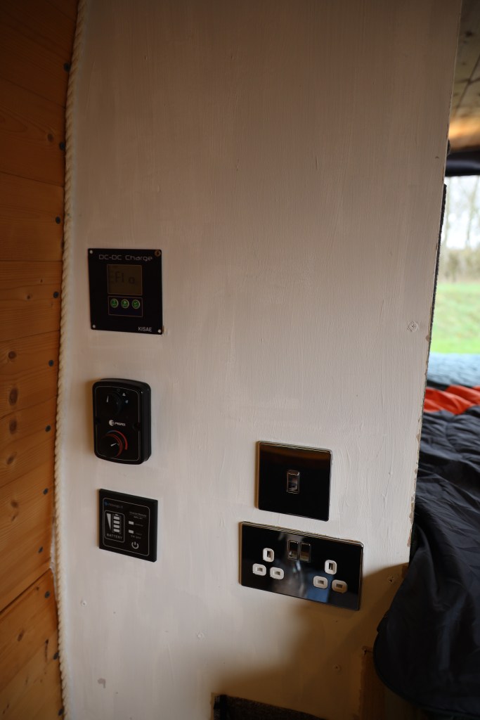 A wall section displaying multiple electrical panels and outlets, including a DC-DC charger, a power control switch, and various power sockets in a cozy indoor setting.
