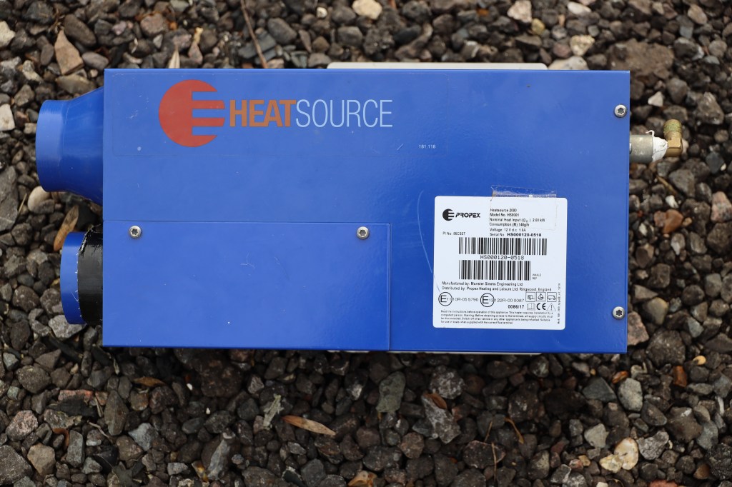 A blue heat source unit labeled 'HEAT SOURCE' with connections and a product information sticker, positioned on gravel.